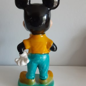 Op de afbeelding: Een vintage Mickey Mouse beeldje, van achteren gezien. De figuur heeft zwarte oren en hoofd, een geel shirt, een turquoise broek en gele schoenen. Het beeldje staat op een groene basis, met een witte handschoen zichtbaar.