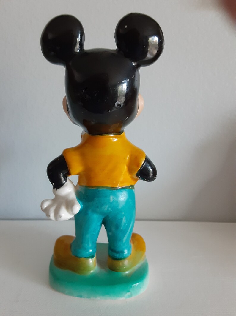Op de afbeelding: Een vintage Mickey Mouse beeldje, van achteren gezien. De figuur heeft zwarte oren en hoofd, een geel shirt, een turquoise broek en gele schoenen. Het beeldje staat op een groene basis, met een witte handschoen zichtbaar.