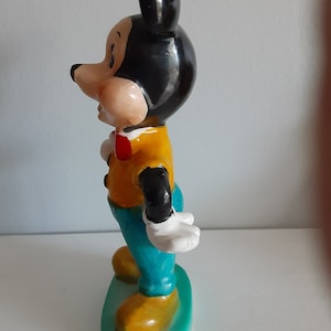 Op de afbeelding: Een vintage Mickey Mouse beeldje. De figuur draagt een geel shirt, een blauwe broek en een rode vlinderdas. De oren en het haar zijn zwart en de schoenen zijn geel. Het beeldje staat op een groene basis.