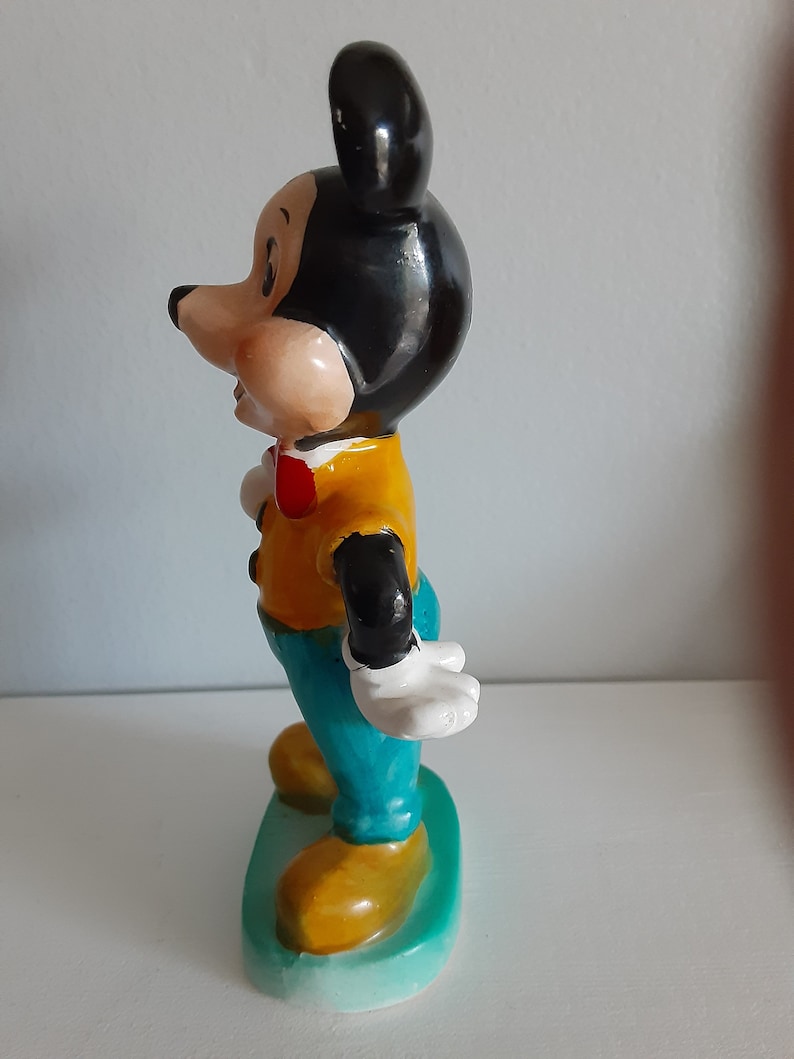 Op de afbeelding: Een vintage Mickey Mouse beeldje. De figuur draagt een geel shirt, een blauwe broek en een rode vlinderdas. De oren en het haar zijn zwart en de schoenen zijn geel. Het beeldje staat op een groene basis.