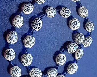 May include: A blue and white ceramic necklace with a knotted cord. The necklace features round beads with a white background and a blue geometric design.