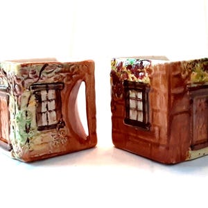 May include: Two ceramic cream pitchers shaped like miniature houses. The pitchers are brown with detailed facades, including windows, doors, and climbing vines. One pitcher has a handle. The design evokes a rustic, cottagecore aesthetic.