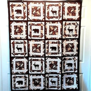 May include: A brown and white quilt with a repeating pattern of squares. Each square features a silhouette of a bear or deer. The quilt has a dark brown border and is hanging on a white door.