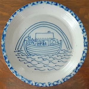 May include: A white ceramic plate with a blue rim and a hand-drawn illustration of Noah's Ark. The image depicts the ark with animals, a rainbow, and water waves. The plate has a rustic, handmade appearance.