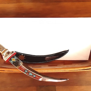 May include: An ornate dagger with a curved blade, featuring a gold-colored handle with intricate patterns and red, blue, and turquoise accents. The dagger rests in a wooden basket with a black sheath and a stack of white paper.