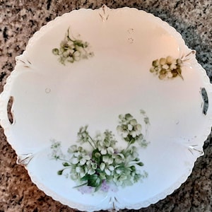 May include: A white porcelain serving plate with a scalloped edge and two handles. The plate is decorated with hand-painted green and purple floral clusters and gold accents. The plate measures approximately 10 inches in diameter.