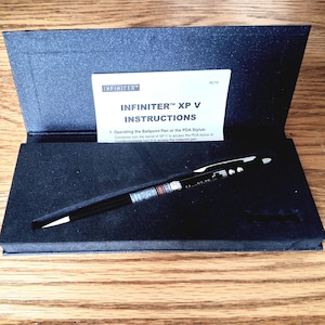 May include: A black Infiniter XP V ballpoint pen and PDA stylus in a black presentation box with an instruction card. The card reads INFINITER XP V INSTRUCTIONS and includes operating steps. The pen features a silver clip and a laser warning label.