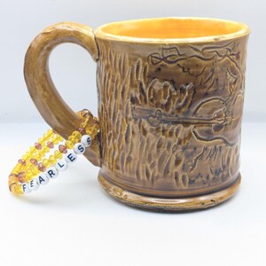 Puede incluir: Una taza de cer&aacute;mica marr&oacute;n con un dise&ntilde;o de corteza de &aacute;rbol y una pulsera de cuentas amarillas y naranjas con la palabra "FEARLESS" escrita en ella.