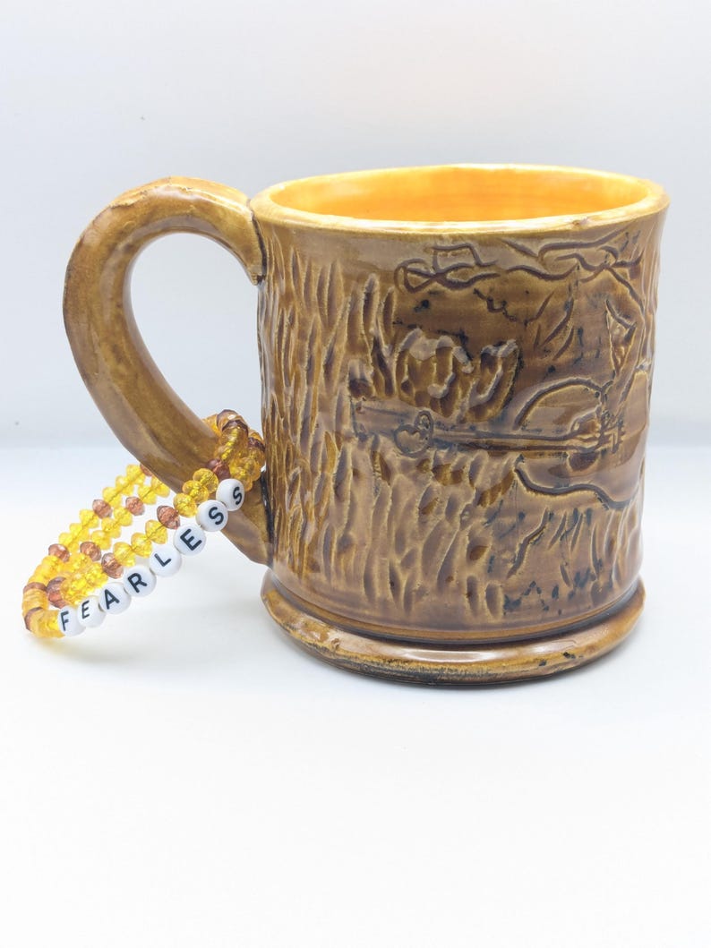 Puede incluir: Una taza de cer&aacute;mica marr&oacute;n con un dise&ntilde;o de corteza de &aacute;rbol y una pulsera de cuentas amarillas y naranjas con la palabra "FEARLESS" escrita en ella.