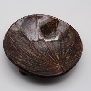 May include: A small, brown ceramic dish with a leaf design. The dish has a glossy finish and is slightly raised on three small feet.