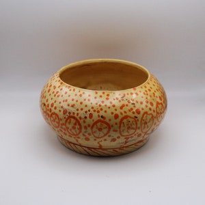 Puede incluir: Un cuenco de cerámica amarillo con un diseño moteado naranja y rojo. El cuenco tiene una forma redondeada y un borde ancho.