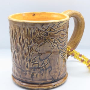 Puede incluir: Una taza de cer&aacute;mica marr&oacute;n con una superficie texturizada y un dise&ntilde;o de una cara de mujer con el pelo suelto. La taza tiene un esmalte amarillo en el interior y un asa marr&oacute;n.