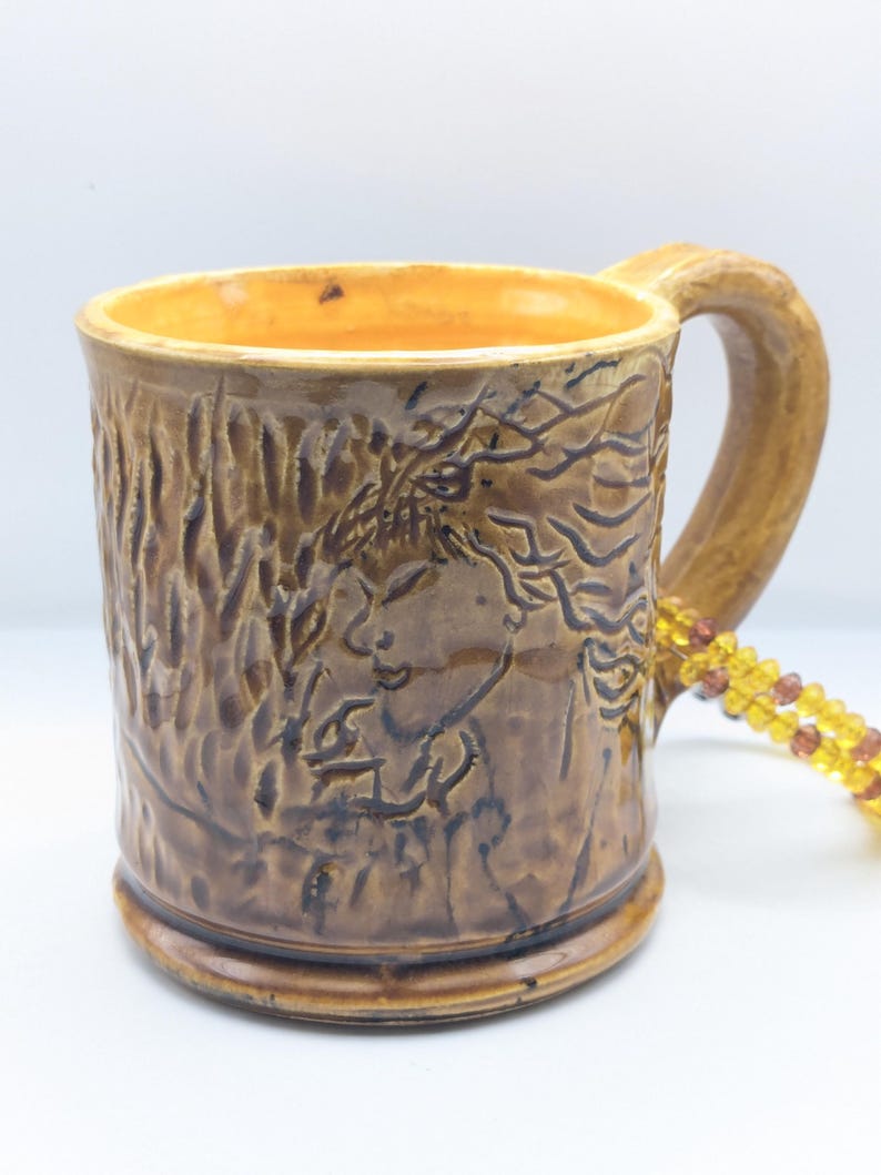 Puede incluir: Una taza de cer&aacute;mica marr&oacute;n con una superficie texturizada y un dise&ntilde;o de una cara de mujer con el pelo suelto. La taza tiene un esmalte amarillo en el interior y un asa marr&oacute;n.