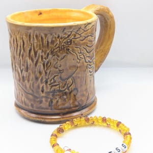 Puede incluir: Una taza de cer&aacute;mica marr&oacute;n con un dise&ntilde;o en relieve de una cara de mujer y una pulsera de cuentas amarillas y marrones con la palabra "FEARLESS" escrita en letras blancas.