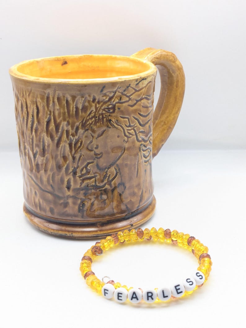 Puede incluir: Una taza de cer&aacute;mica marr&oacute;n con un dise&ntilde;o en relieve de una cara de mujer y una pulsera de cuentas amarillas y marrones con la palabra "FEARLESS" escrita en letras blancas.