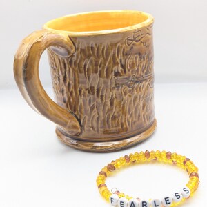 Puede incluir: Una taza de cer&aacute;mica marr&oacute;n con una superficie texturizada y un asa. La taza tiene un dise&ntilde;o de un oso. Una pulsera de cuentas amarillas con la palabra "FEARLESS" escrita en letras blancas.