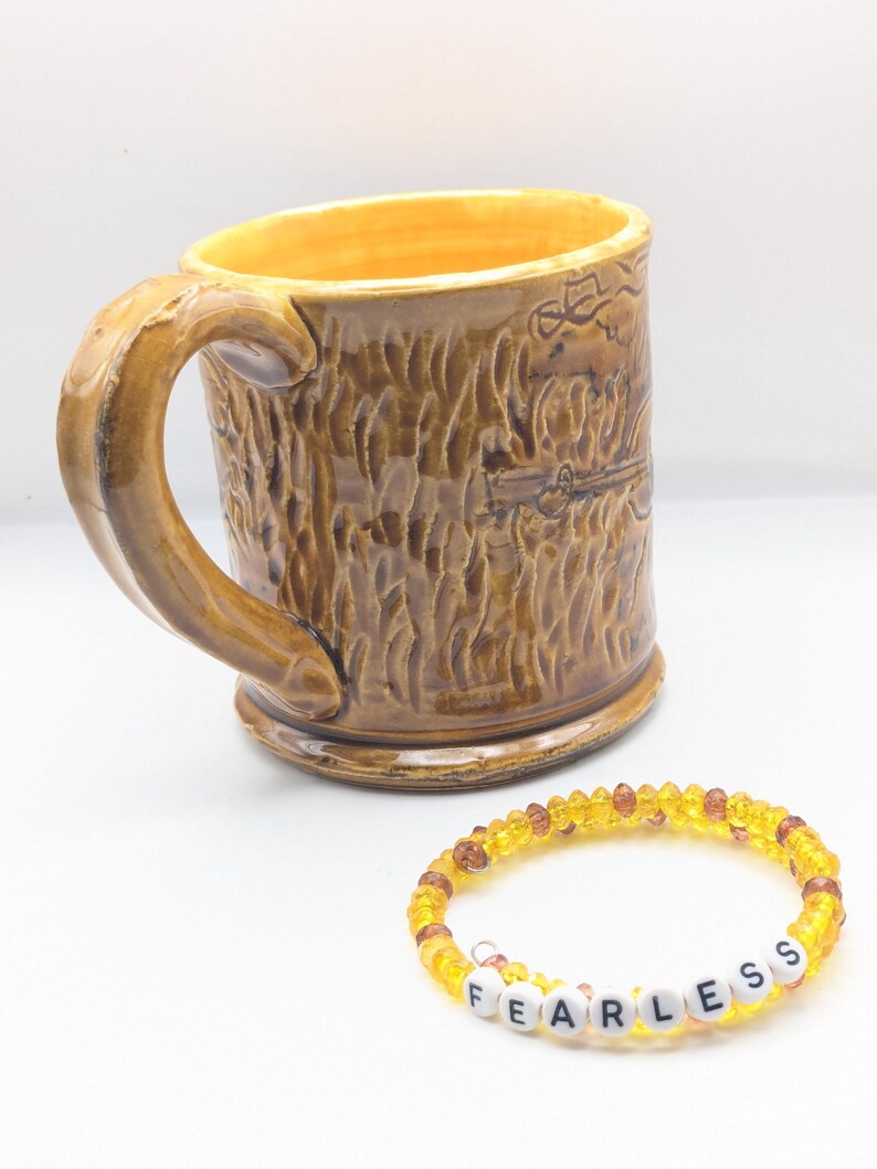 Puede incluir: Una taza de cer&aacute;mica marr&oacute;n con una superficie texturizada y un asa. La taza tiene un dise&ntilde;o de un oso. Una pulsera de cuentas amarillas con la palabra "FEARLESS" escrita en letras blancas.