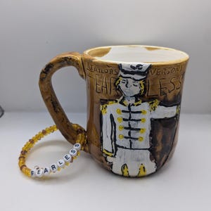 Peut inclure: Une tasse en céramique brune avec un motif blanc représentant une personne en uniforme militaire. La tasse porte les mots "Taylor's Version" et "Fearless". Un bracelet en perles brunes avec des perles blanches épellant "Fearless" est au premier plan.