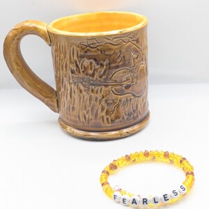 Puede incluir: Una taza de cer&aacute;mica marr&oacute;n con un dise&ntilde;o de &aacute;rbol y guitarra. Una pulsera de cuentas amarillas con la palabra "FEARLESS" escrita en letras blancas.