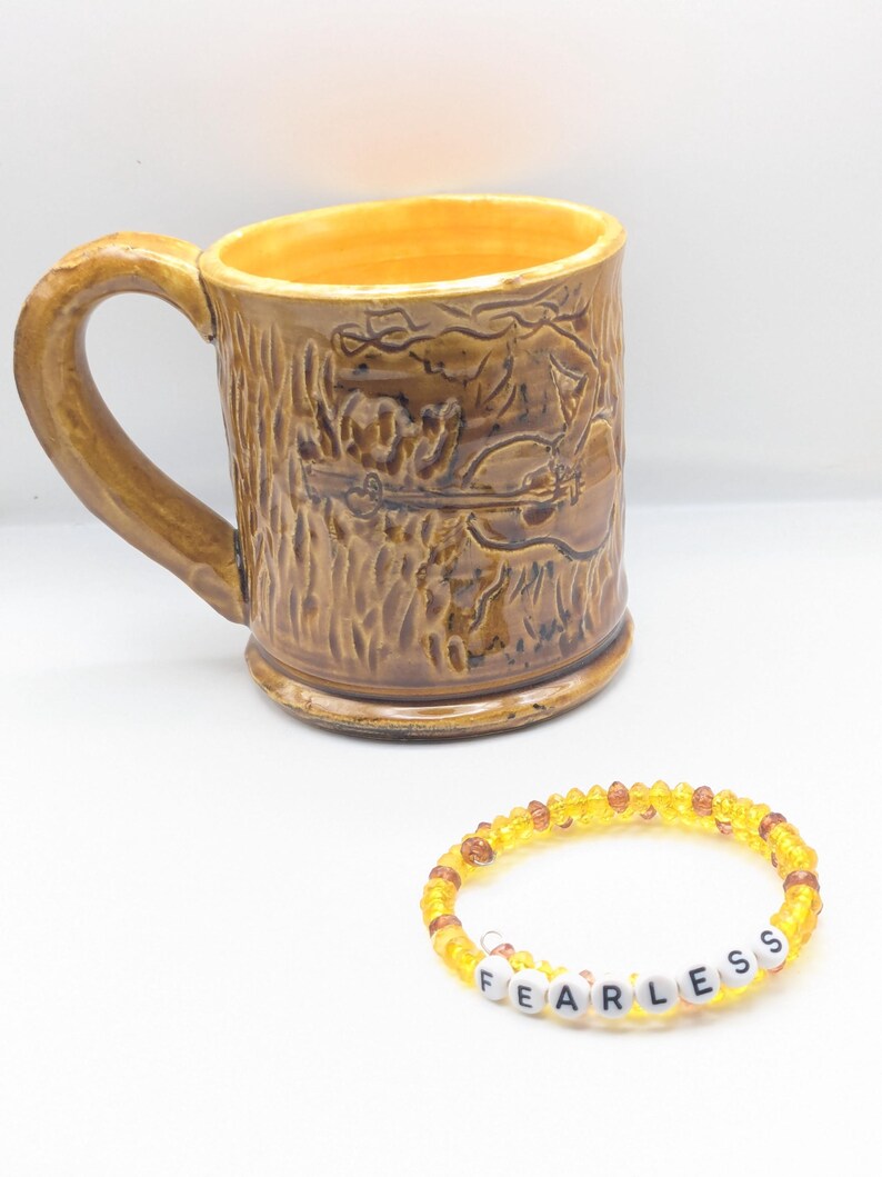 Puede incluir: Una taza de cer&aacute;mica marr&oacute;n con un dise&ntilde;o de &aacute;rbol y guitarra. Una pulsera de cuentas amarillas con la palabra "FEARLESS" escrita en letras blancas.