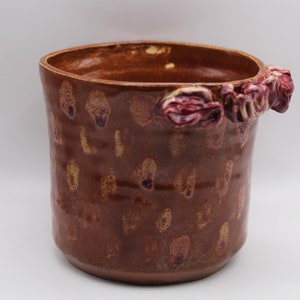 May include: A brown ceramic planter with a textured surface and a raised design of red flowers around the rim.