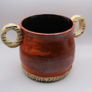 May include: A brown ceramic mug with two white handles decorated with black and orange stripes. The mug has a black interior and a brown glaze.