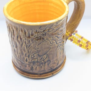 Puede incluir: Una taza de cer&aacute;mica marr&oacute;n con una superficie texturizada y un dise&ntilde;o de &aacute;rbol. La taza tiene un interior amarillo y un asa. Un collar de cuentas est&aacute; en el fondo.