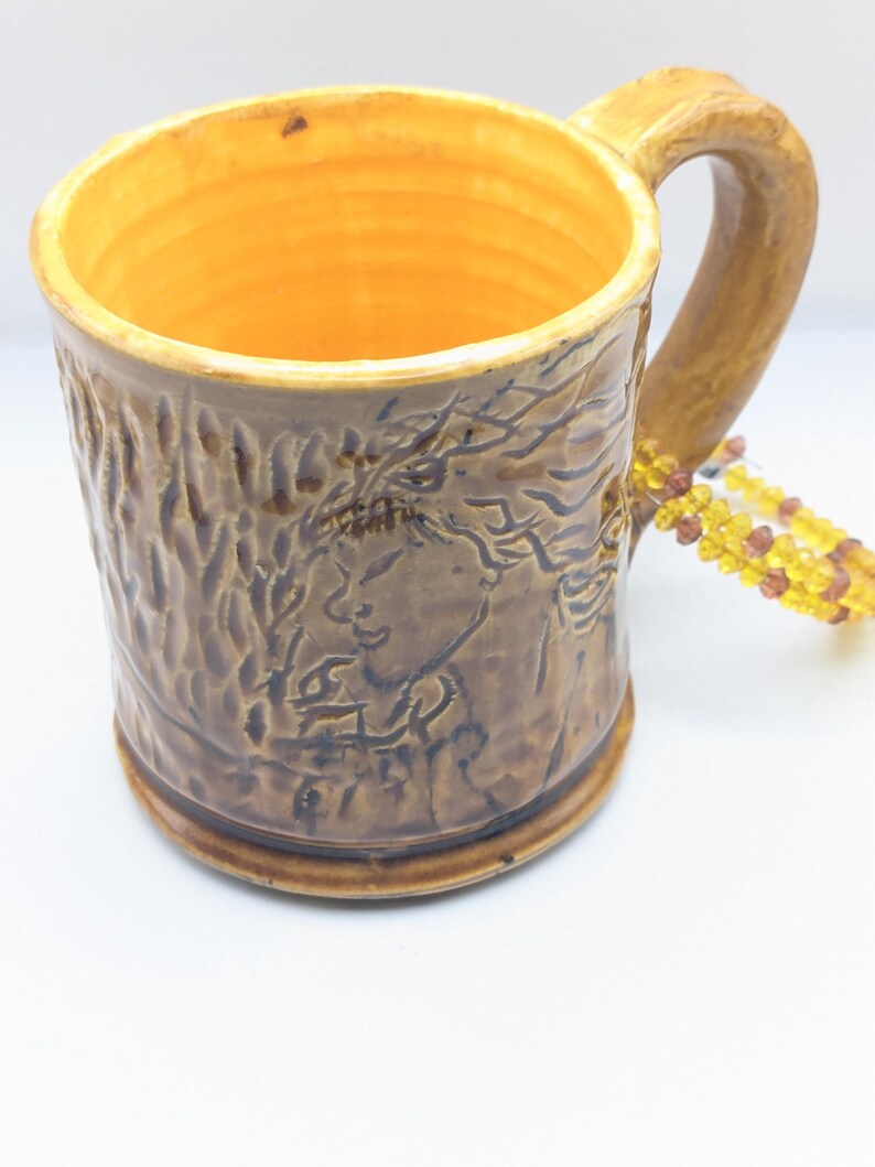 Puede incluir: Una taza de cer&aacute;mica marr&oacute;n con una superficie texturizada y un dise&ntilde;o de &aacute;rbol. La taza tiene un interior amarillo y un asa. Un collar de cuentas est&aacute; en el fondo.
