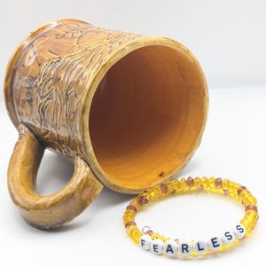 Puede incluir: Una taza de cer&aacute;mica marr&oacute;n con un dise&ntilde;o texturizado y una pulsera de cuentas amarillas con la palabra "FEARLESS" escrita en cuentas blancas.
