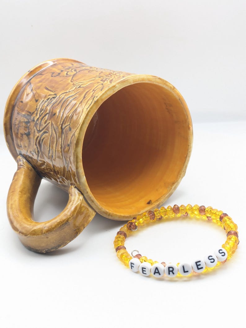 Puede incluir: Una taza de cer&aacute;mica marr&oacute;n con un dise&ntilde;o texturizado y una pulsera de cuentas amarillas con la palabra "FEARLESS" escrita en cuentas blancas.