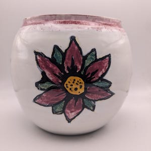 Puede incluir: Maceta de cerámica blanca con una flor rosa pintada a mano con un centro amarillo y hojas verdes.