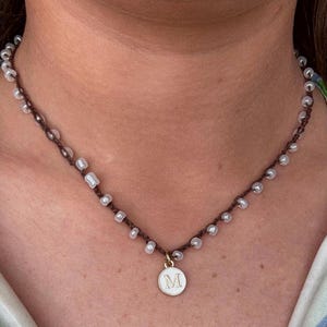 Puede incluir: Collar hecho a mano con un cordón marrón oscuro y pequeñas cuentas blancas. El collar tiene un colgante dorado con una cara de esmalte blanco y la letra "M". Estilo gargantilla.