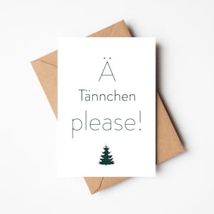 Könnte beinhalten: Eine weiße Grußkarte mit dem Text "Ä Tännchen please!" in Dunkelgrün und einer kleinen Weihnachtsbaum-Illustration. Die Karte liegt auf einem braunen Umschlag vor weißem Hintergrund.