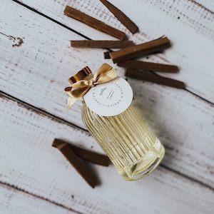 May include: A clear glass bottle with a gold lid and a white label that reads "D'AMBIANCE  CREATIONS  PARFUM  Thistle  Dark Amber & Sandalwood". The bottle is sitting on a white wooden surface with brown wooden sticks scattered around it.