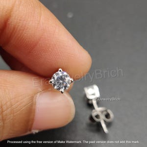 May include: Close-up of a silver stud earring featuring a clear, round gemstone. The earring is held between fingers, with another earring in the background. The earring has a post and butterfly backing.