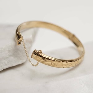 May include: A gold bangle bracelet with a delicate chain attached. The bracelet has an engraved floral design.