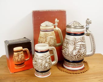 May include: Three ceramic beer steins with images of vintage cars. The largest stein has a blue and white design with the text "1911 Ford Model T" and a silver lid. The smallest stein has a blue and white design with the text "1911 Ford Model T". The middle stein has a blue and white design with the text "1911 Ford Model T".