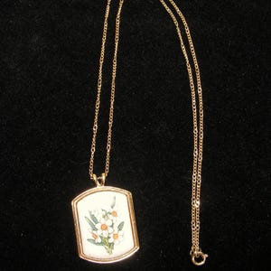 May include: A gold-coloured necklace with a rectangular pendant. The pendant displays a floral design on a white background, with white and orange flowers and green leaves. The chain is fine and has a simple clasp.