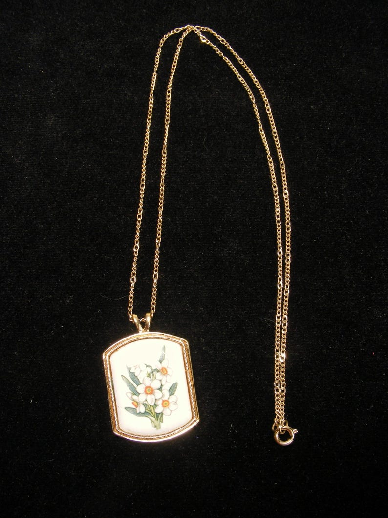 May include: A gold-coloured necklace with a rectangular pendant. The pendant displays a floral design on a white background, with white and orange flowers and green leaves. The chain is fine and has a simple clasp.
