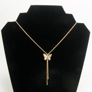 May include: Gold-coloured butterfly pendant necklace. The butterfly pendant is encrusted with small, clear crystals. The necklace has a delicate chain and a dangling tassel design. The necklace is displayed on a black jewellery stand.