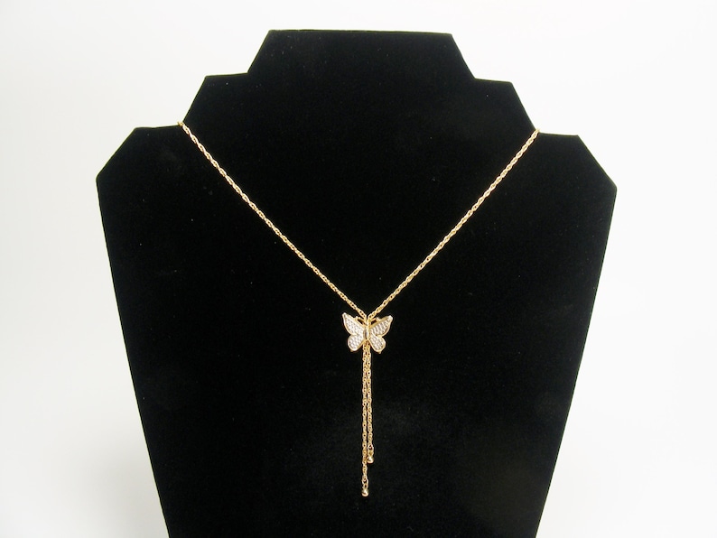 May include: Gold-coloured butterfly pendant necklace. The butterfly pendant is encrusted with small, clear crystals. The necklace has a delicate chain and a dangling tassel design. The necklace is displayed on a black jewellery stand.