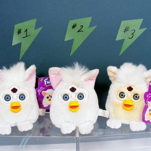 May include: Three white Furby toys with blue eyes, yellow beaks, and pink or tan ears are displayed. Each has a tag. Green lightning bolt-shaped signs above them are labeled #1, #2, and #3. The toys are on a clear shelf against a dark blue background.