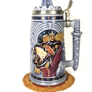 May include: A ceramic beer stein featuring a relief of a crowned monarch in armor with a red cape. The lid is topped with a miniature castle sculpture and includes a metal thumb lever. The handle is textured with a cross pattern in blue and grey.