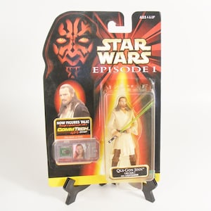 May include: Star Wars Episode I action figure packaging featuring Qui-Gon Jinn with lightsaber. The packaging has a red and yellow background with the Star Wars logo and the words "Now figures talk!" and "CommTech Chip".