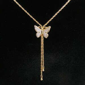 May include: A gold butterfly pendant necklace with a fine chain. The butterfly has a textured, gem-like appearance on its wings. Two strands of chain hang below the butterfly, each ending with a small, round charm. The background is black.