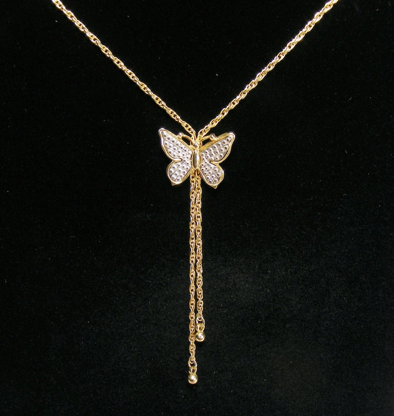 May include: A gold butterfly pendant necklace with a fine chain. The butterfly has a textured, gem-like appearance on its wings. Two strands of chain hang below the butterfly, each ending with a small, round charm. The background is black.