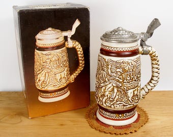 May include: A ceramic beer stein with a cowboy roping a steer design. The stein has a brown and white rope handle and a metal lid with a hinged thumb lift. The stein is sitting on a crocheted doily. The box says "Roping Chuck Wagon"