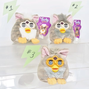 May include: Three Furby Buddies plush toys. The toys have gray and white fur, pink ears, and yellow beaks. Each Furby has a tag attached. The toys are displayed on a clear shelf.