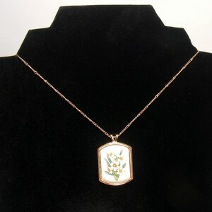 May include: A gold-coloured necklace with a rectangular pendant featuring a floral design. The pendant has a white background with yellow and green flowers. The necklace is displayed on a black velvet stand.