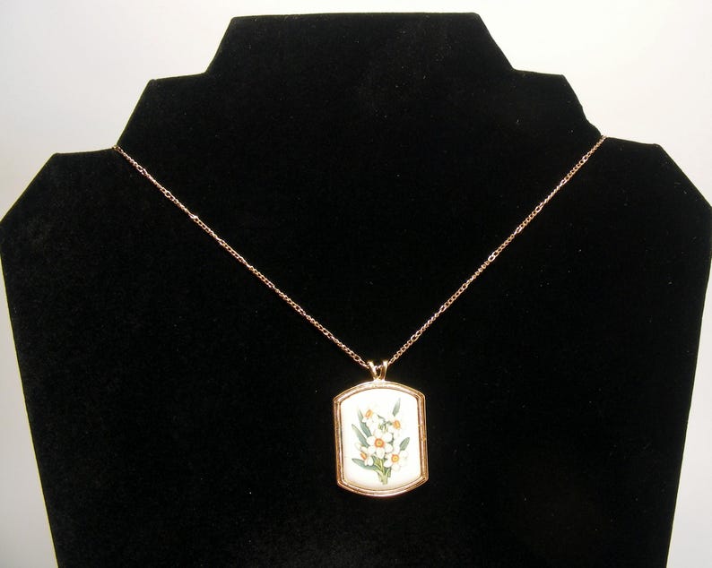 May include: A gold-coloured necklace with a rectangular pendant featuring a floral design. The pendant has a white background with yellow and green flowers. The necklace is displayed on a black velvet stand.